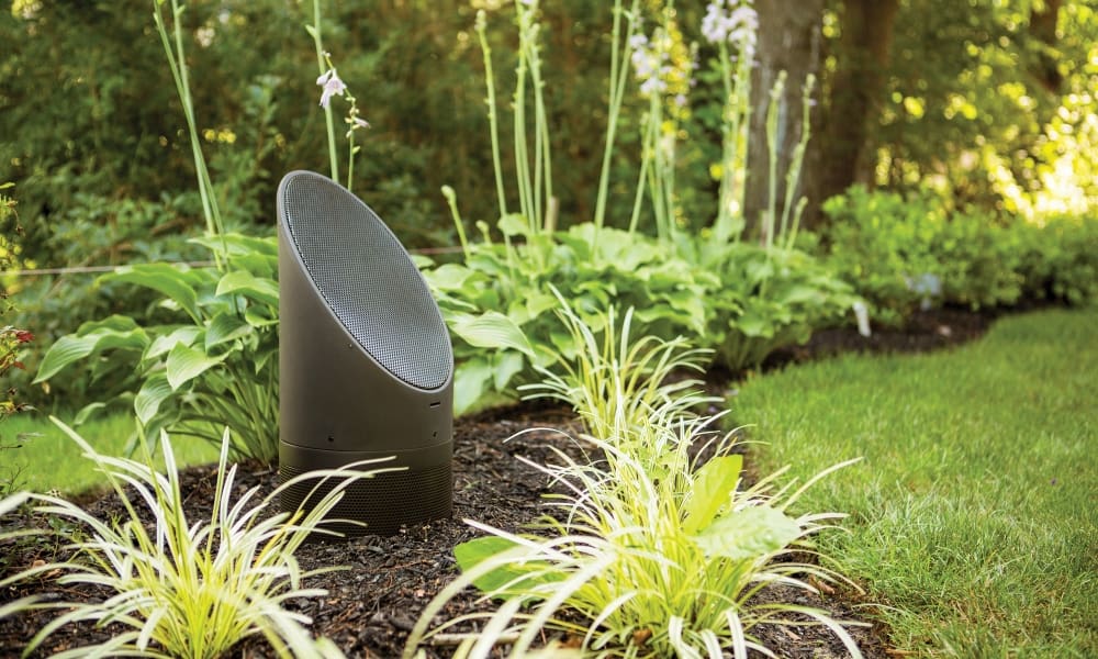 A camouflaged outdoor speaker designed to look like a natural rock, placed among plants and mulch in a landscaped garden setting.